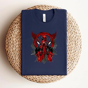 K&ouml;nnte beinhalten: Marineblaues T-Shirt mit einem Deadpool-Motiv. Das Design zeigt Deadpool in einem rot-schwarzen Anzug, der zwei Katanas h&auml;lt, mit einem roten Kreissymbol und Fl&uuml;geln. Das T-Shirt ist auf einer gewebten Oberfl&auml;che gefaltet.