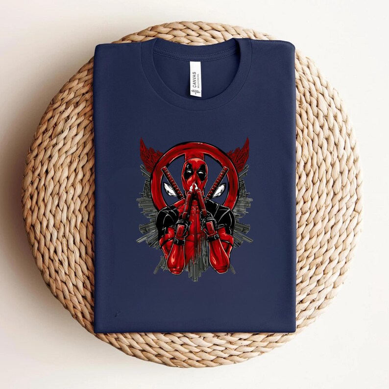 K&ouml;nnte beinhalten: Marineblaues T-Shirt mit einem Deadpool-Motiv. Das Design zeigt Deadpool in einem rot-schwarzen Anzug, der zwei Katanas h&auml;lt, mit einem roten Kreissymbol und Fl&uuml;geln. Das T-Shirt ist auf einer gewebten Oberfl&auml;che gefaltet.
