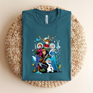 Op de afbeelding: Tealkleurig T-shirt met een kleurrijke afbeelding van geanimeerde personages in ruimte-thema outfits. Het ontwerp bevat een rendier in een ruimtepak, een sneeuwpop en andere personages die futuristische wapens vasthouden.