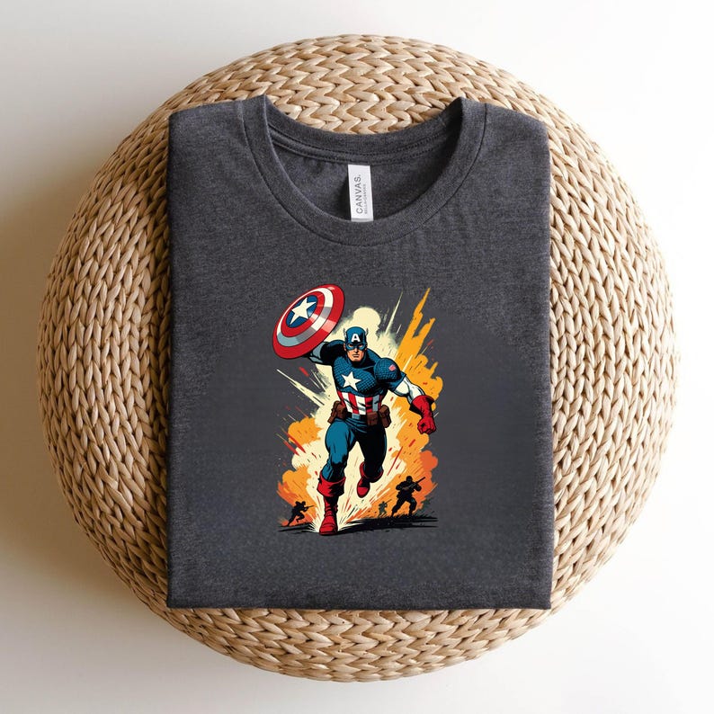 May include: Dark gray t-shirt featuring a colorful illustration of Captain America with his shield, running in front of an explosion. The design includes the iconic star-spangled suit and shield. The shirt is displayed on a woven surface.