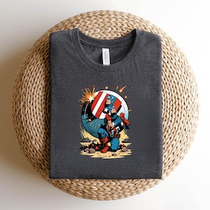 Puede incluir: Camiseta gris oscuro con un dise&ntilde;o de Capit&aacute;n Am&eacute;rica. El dise&ntilde;o muestra al Capit&aacute;n Am&eacute;rica arrodillado con su escudo, en colores rojo, blanco y azul. La camiseta est&aacute; hecha de un material suave.