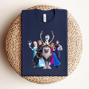 Op de afbeelding: Marineblauw T-shirt met een afbeelding van personages uit de animatiefilm Frozen. Het ontwerp omvat Elsa, Anna, Kristoff, Sven, Olaf en Hans. Het shirt is gemaakt van een zacht materiaal en wordt gepresenteerd op een geweven oppervlak.