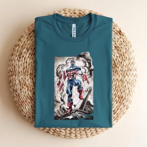 May include: Teal-colored t-shirt featuring a graphic of Captain America in a heroic pose, holding his shield amidst a smoky background. The illustration is detailed with red, white, and blue colors, capturing the iconic superhero's attire. The shirt is folded on a woven surface.