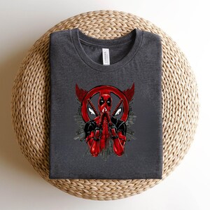 K&ouml;nnte beinhalten: Dunkelgraues T-Shirt mit einem Deadpool-Motiv. Das Design zeigt Deadpool in einem rot-schwarzen Anzug, der zwei Katanas h&auml;lt, vor einem roten Emblem mit Fl&uuml;geln und einer Stadtlandschaft. Das T-Shirt ist aus weichem Material.