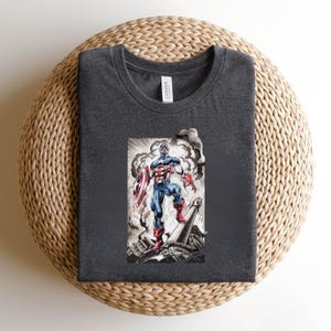 May include: Dark gray t-shirt featuring a graphic of Captain America with his shield, standing amidst a smoky, war-torn scene. The illustration is in shades of red, white, blue, and gray. The shirt is made of a soft material.
