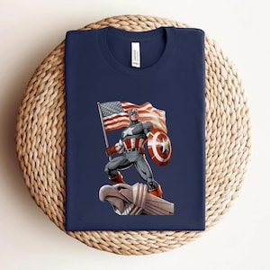 Pode incluir: Camiseta azul marinho com um desenho do Capit&atilde;o Am&eacute;rica segurando uma bandeira e um escudo americanos. A ilustra&ccedil;&atilde;o &eacute; em estilo de quadrinhos, com cores vibrantes e linhas arrojadas. A camiseta est&aacute; dobrada sobre uma superf&iacute;cie tecida.