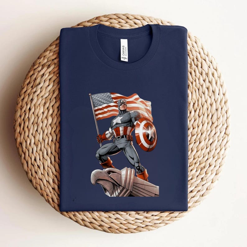 Pode incluir: Camiseta azul marinho com um desenho do Capit&atilde;o Am&eacute;rica segurando uma bandeira e um escudo americanos. A ilustra&ccedil;&atilde;o &eacute; em estilo de quadrinhos, com cores vibrantes e linhas arrojadas. A camiseta est&aacute; dobrada sobre uma superf&iacute;cie tecida.