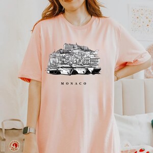 K&ouml;nnte beinhalten: Hellrosa T-Shirt mit einer Schwarz-Wei&szlig;-Illustration der Skyline von Monaco und Yachten. Das Wort "MONACO" ist darunter gedruckt. Das Shirt hat kurze &Auml;rmel und einen Rundhalsausschnitt.