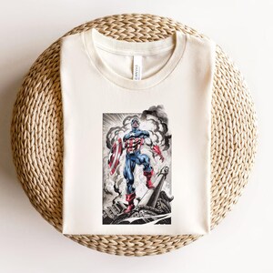 May include: Cream-colored t-shirt featuring a comic book illustration of Captain America. The artwork depicts Captain America in his iconic red, white, and blue suit, holding his shield, standing on a destroyed structure with smoke in the background.