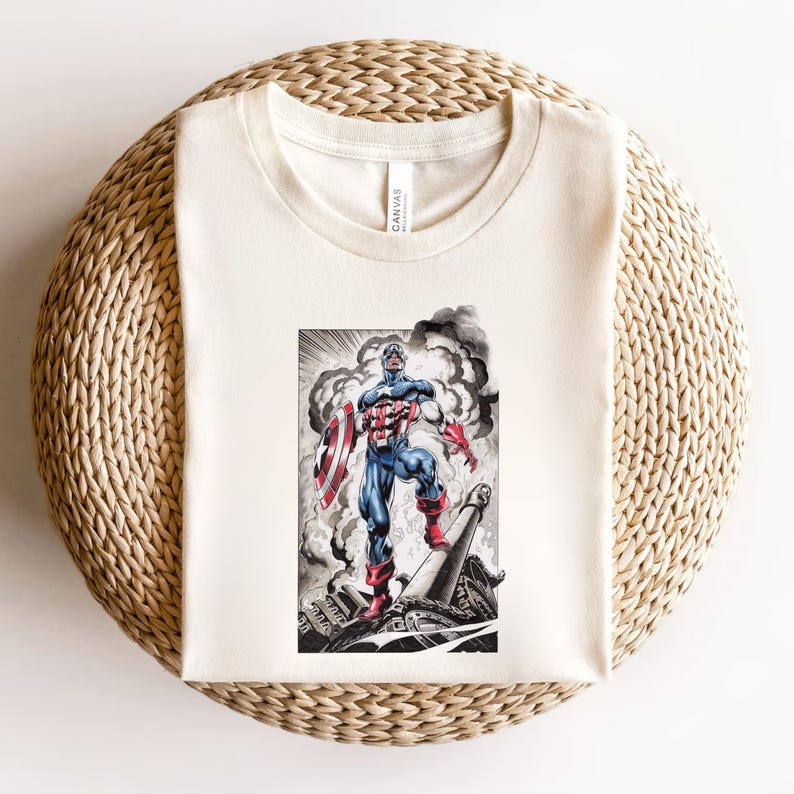 May include: Cream-colored t-shirt featuring a comic book illustration of Captain America. The artwork depicts Captain America in his iconic red, white, and blue suit, holding his shield, standing on a destroyed structure with smoke in the background.