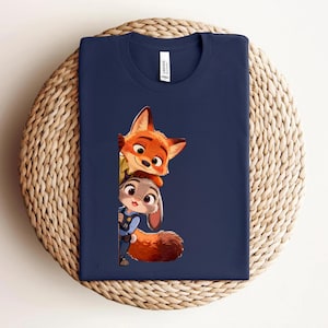 May include: A navy blue crew neck t-shirt featuring a graphic print of a cartoon fox and a cartoon rabbit peeking out from behind a vertical edge. The fox is orange with large eyes, and the rabbit is grey with a blue uniform and a bushy tail.
