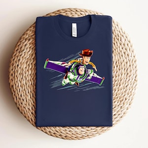 May include: Navy blue t-shirt featuring a graphic of Woody and Buzz Lightyear flying together. The design shows the characters in flight with a purple and green wing design. The shirt is folded on a woven, circular surface.