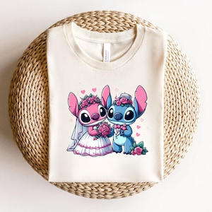 May include: A cream-colored t-shirt featuring a graphic of two cartoon characters dressed for a wedding. One character is pink with a white veil and dress, holding a bouquet of pink roses. The other is blue, wearing a bowtie and holding a rose.