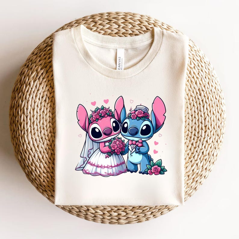 May include: A cream-colored t-shirt featuring a graphic of two cartoon characters dressed for a wedding. One character is pink with a white veil and dress, holding a bouquet of pink roses. The other is blue, wearing a bowtie and holding a rose.