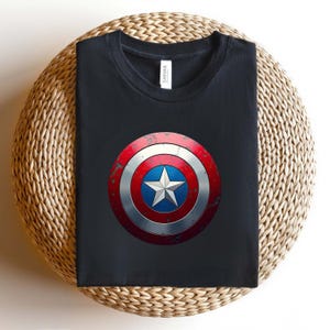 May include: A black crew neck t-shirt featuring a centred graphic of a circular shield with a distressed red, white, and blue finish. The shield design includes a prominent white five-pointed star set against a blue circular background.