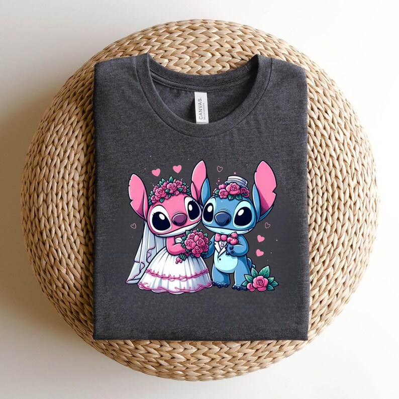 May include: A dark heather gray crew neck t-shirt featuring a graphic of two blue and pink alien characters dressed for a wedding. The pink character wears a white veil and dress, while the blue character wears a tuxedo, both adorned with pink roses.