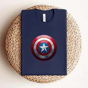 May include: A dark navy blue crew neck t-shirt featuring a centred graphic of a circular shield with a distressed red, white, and blue finish and a prominent white star in the middle. The shirt has a white brand label inside the collar area.