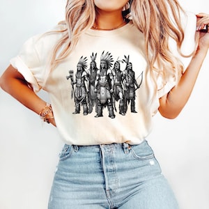 May include: Cream-colored t-shirt featuring a black and white illustration of six Native American figures in traditional attire. The figures are depicted with headdresses, bows, and arrows, and other tribal accessories. The shirt is a casual wear item.