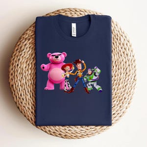 May include: Navy blue t-shirt featuring a colorful graphic of characters from the animated movie Toy Story. The design includes a pink bear, a cowgirl, a cowboy, and a space ranger. The shirt is displayed on a woven surface.