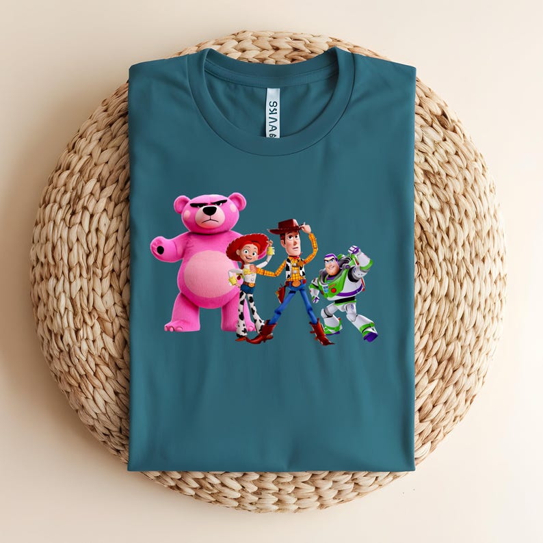 May include: Teal t-shirt featuring a graphic of characters from the animated movie Toy Story. The design includes a pink bear, Woody, Jessie, and Buzz Lightyear. The shirt is folded on a woven mat.