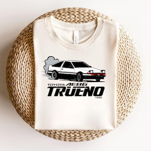 May include: Cream-colored t-shirt featuring a black and white graphic of a Toyota AE86 Trueno car with smoke trails. The text "TOYOTA AE86 TRUENO 1985" is printed below the car in a bold, black font. The shirt is displayed on a woven surface.