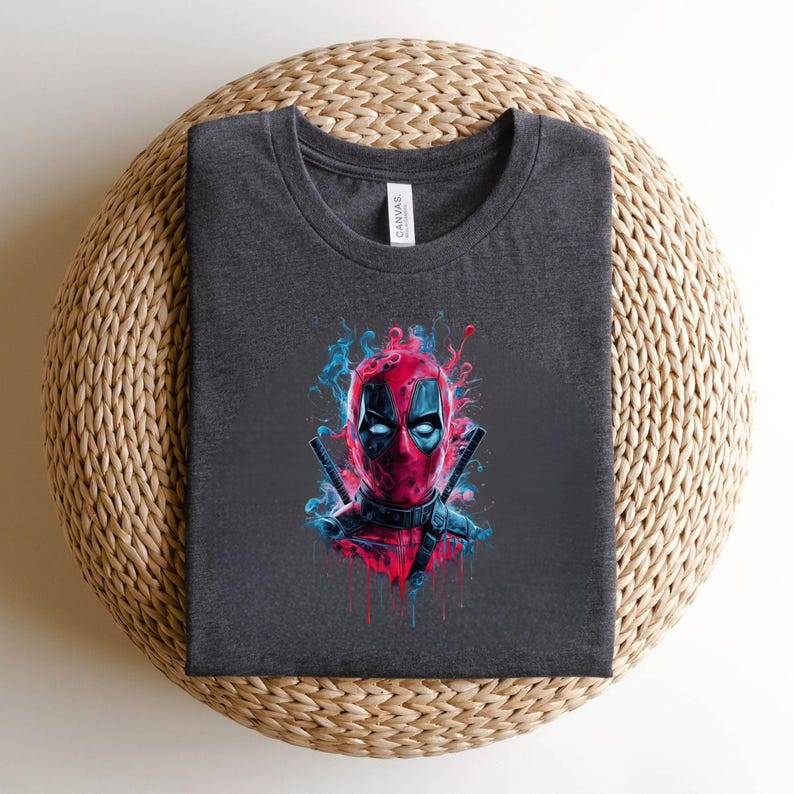 May include: Dark gray t-shirt featuring a colorful graphic of Deadpool's face. The design includes red and blue accents, with two swords behind the character. The shirt is made of a soft material.