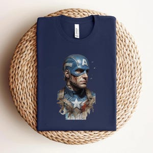 May include: Navy blue t-shirt featuring a Captain America graphic. The graphic depicts Captain America's face and helmet with the letter "A" on the helmet. The shirt is folded on a woven surface.