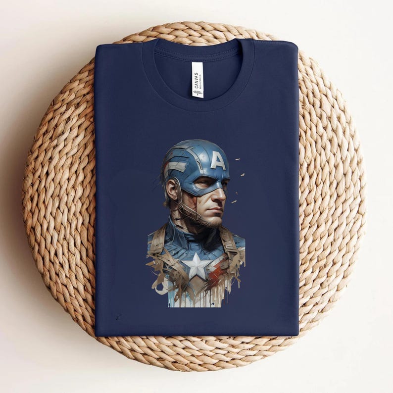 May include: Navy blue t-shirt featuring a Captain America graphic. The graphic depicts Captain America's face and helmet with the letter "A" on the helmet. The shirt is folded on a woven surface.