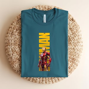 May include: Teal t-shirt featuring a graphic of Iron Man in his red and gold armor. The word "IRON MAN" is printed vertically in yellow. The shirt is displayed on a woven, circular surface.