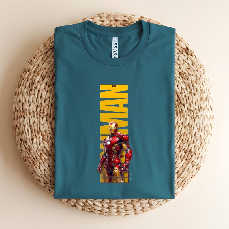 May include: Teal t-shirt featuring a graphic of Iron Man in his red and gold armor. The word "IRON MAN" is printed vertically in yellow. The shirt is displayed on a woven, circular surface.