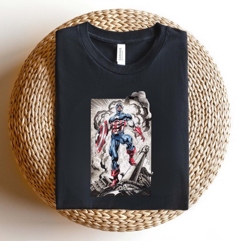 May include: Black t-shirt featuring a graphic of Captain America in a heroic pose, holding his shield. The illustration is in a comic book style with a detailed background of smoke and debris. The shirt is displayed on a woven, circular surface.