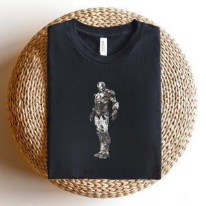 May include: Black t-shirt featuring a detailed illustration of a robotic figure. The artwork is in shades of gray, white, and gold, with blue accents. The shirt is folded and placed on a woven, circular surface.