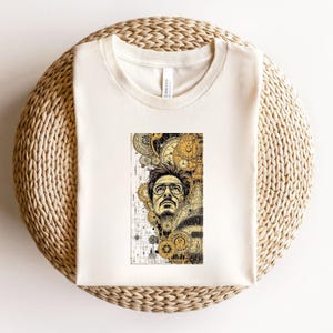 Pode incluir: Camiseta creme com uma ilustra&ccedil;&atilde;o detalhada de um homem com &oacute;culos, integrada com mecanismos de rel&oacute;gios e engrenagens. O design &eacute; em tons de ouro, preto e branco. A camiseta &eacute; feita de um material macio.