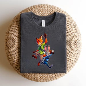 Puede incluir: Una camiseta de cuello redondo gris carb&oacute;n oscuro con un estampado gr&aacute;fico de un zorro de dibujos animados con camisa verde y un conejo de dibujos animados con uniforme de polic&iacute;a azul. El zorro salta con un brazo alzado y el conejo corre.