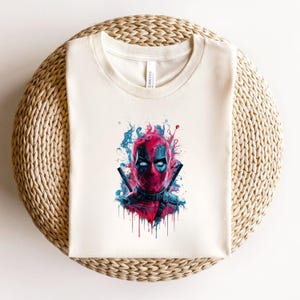 May include: Cream-colored t-shirt featuring a vibrant graphic of Deadpool's face. The artwork showcases a red mask with black eye cutouts, two katanas, and blue and red paint splatter effects. The shirt is displayed on a woven surface.