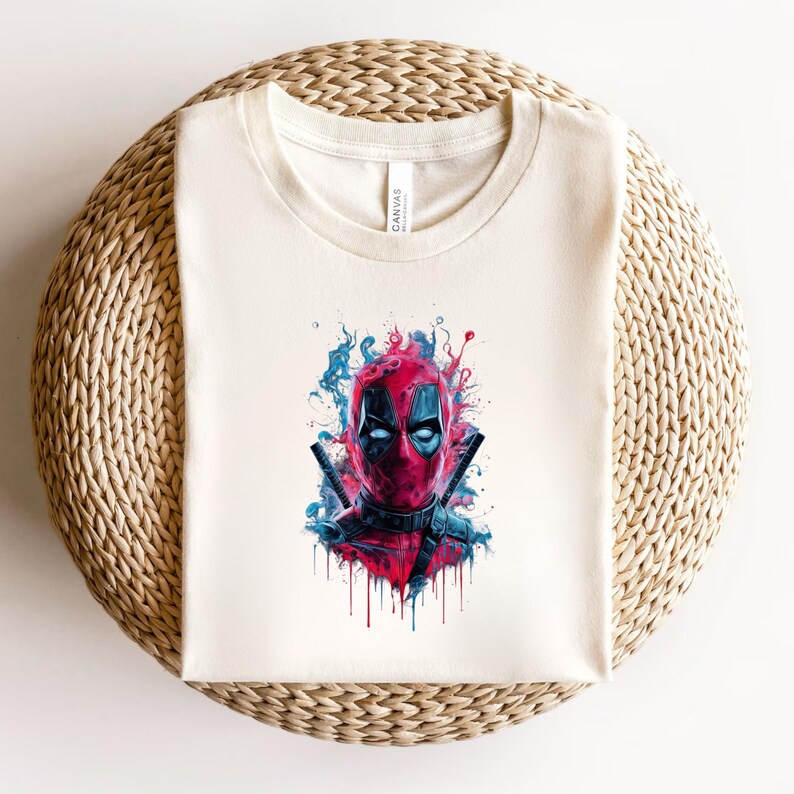 May include: Cream-colored t-shirt featuring a vibrant graphic of Deadpool's face. The artwork showcases a red mask with black eye cutouts, two katanas, and blue and red paint splatter effects. The shirt is displayed on a woven surface.