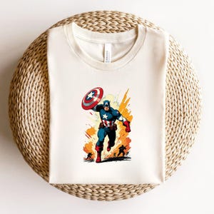 May include: Cream-colored t-shirt featuring a vibrant illustration of Captain America in action. The graphic depicts Captain America in his iconic blue, red, and white suit, holding his shield. The design includes dynamic action lines and silhouettes of other figures.