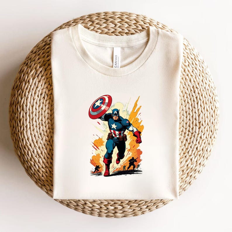 May include: Cream-colored t-shirt featuring a vibrant illustration of Captain America in action. The graphic depicts Captain America in his iconic blue, red, and white suit, holding his shield. The design includes dynamic action lines and silhouettes of other figures.