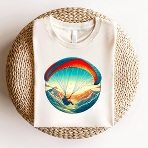 May include: A cream-colored t-shirt featuring a circular graphic print of a paraglider soaring over snow-capped mountain peaks. The paraglider canopy is colored in vibrant shades of red, orange, and blue, set against a bright, sunny sky.