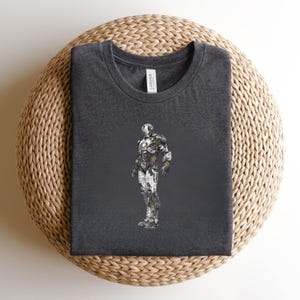 May include: Dark gray t-shirt featuring a detailed illustration of a robotic figure. The design incorporates metallic silver, gold, and blue accents, creating a textured appearance. The shirt is displayed on a woven, circular surface.