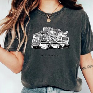 K&ouml;nnte beinhalten: Dunkelgraues T-Shirt mit einer Schwarz-Wei&szlig;-Illustration der Skyline von Monaco und Yachten. Das Wort "MONACO" ist darunter gedruckt. Das T-Shirt hat einen Rundhalsausschnitt und kurze &Auml;rmel.