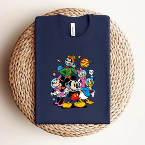 May include: Navy blue t-shirt featuring a colorful graphic of Mickey Mouse, Minnie Mouse, Donald Duck, and Goofy in space-themed attire. The design includes planets, stars, and a rocket ship. The shirt is displayed on a woven surface.