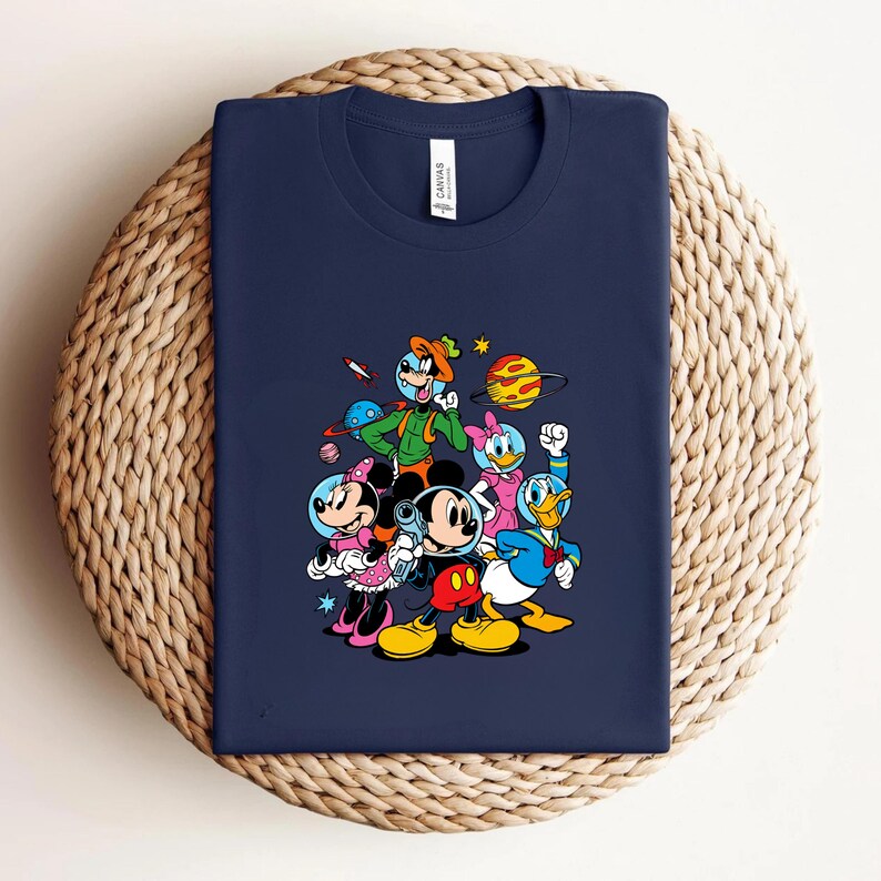 May include: Navy blue t-shirt featuring a colorful graphic of Mickey Mouse, Minnie Mouse, Donald Duck, and Goofy in space-themed attire. The design includes planets, stars, and a rocket ship. The shirt is displayed on a woven surface.
