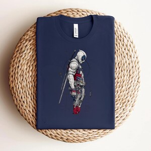 Puede incluir: Una camiseta azul marino con una ilustraci&oacute;n gr&aacute;fica de un personaje que lleva una m&aacute;scara y un traje blancos con detalles en rojo, portando una espada en la espalda. El personaje aparece de perfil con equipo t&aacute;ctico y l&iacute;neas t&eacute;cnicas.