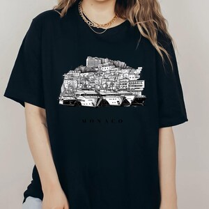 K&ouml;nnte beinhalten: Schwarzes T-Shirt mit einer schwarz-wei&szlig;en Illustration der Skyline von Monaco und Yachten. Das Wort "MONACO" ist darunter gedruckt. Das T-Shirt hat einen Rundhalsausschnitt und ist aus weichem Material.