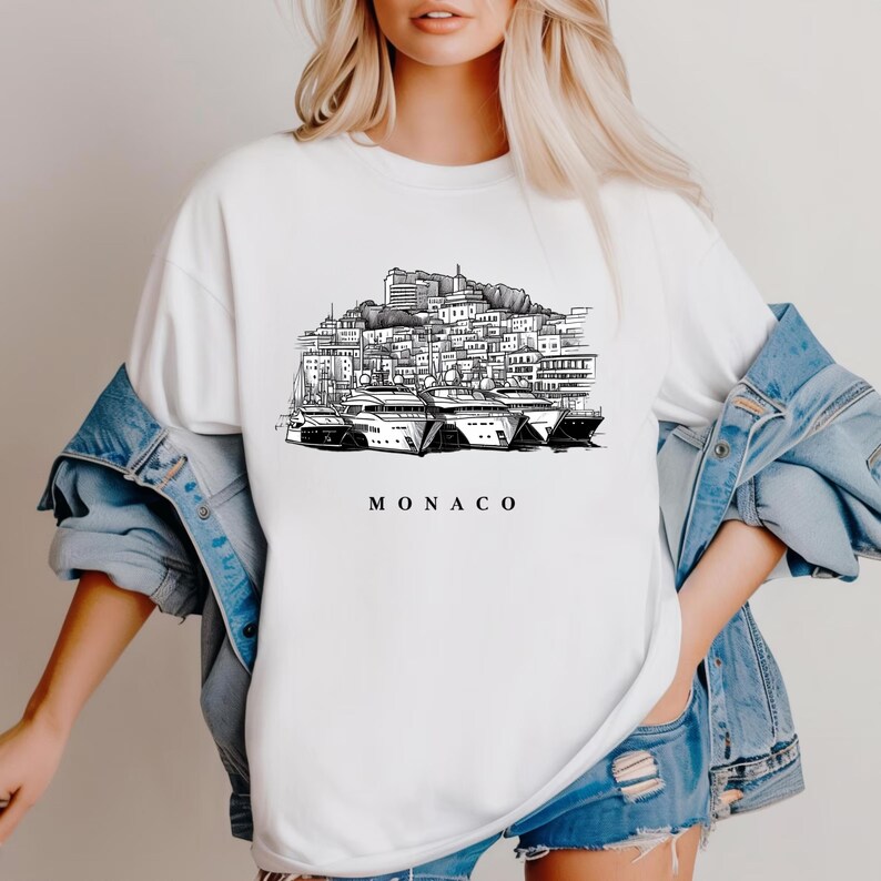 K&ouml;nnte beinhalten: Wei&szlig;es T-Shirt mit einer Schwarz-Wei&szlig;-Illustration der Skyline von Monaco und Yachten. Das Wort "MONACO" ist darunter gedruckt. Das Shirt wird mit einer Jeansjacke und Shorts getragen.