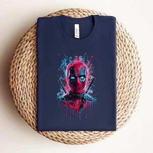 May include: Navy blue t-shirt featuring a vibrant graphic of Deadpool's face with red and blue accents. The design includes two katanas behind the character's head. The shirt is folded on a woven surface.