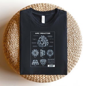 Puede incluir: Camiseta negra con un diagrama detallado de un Reactor Arc, con el texto "ARC REACTOR". El diagrama incluye varios componentes y etiquetas. La camiseta tambi&eacute;n tiene el logo de Stark Industries.