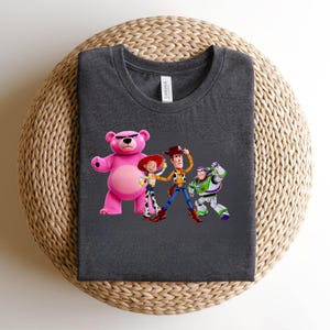 May include: Dark gray t-shirt featuring a graphic of characters from the animated film Toy Story. The design includes a pink bear, Jessie, Woody, and Buzz Lightyear. The shirt is made of a soft material and has a crew neck.