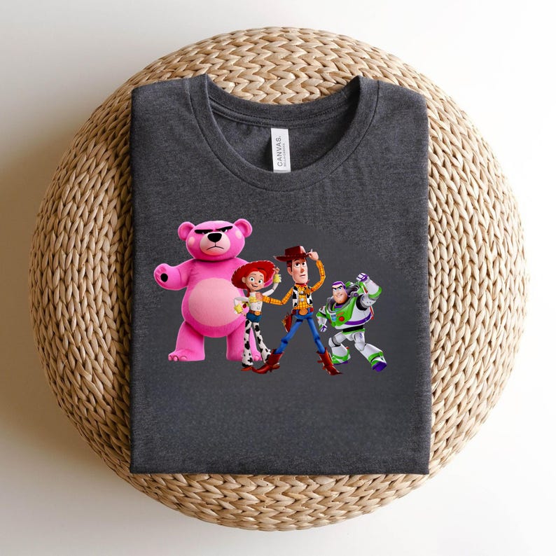 May include: Dark gray t-shirt featuring a graphic of characters from the animated film Toy Story. The design includes a pink bear, Jessie, Woody, and Buzz Lightyear. The shirt is made of a soft material and has a crew neck.